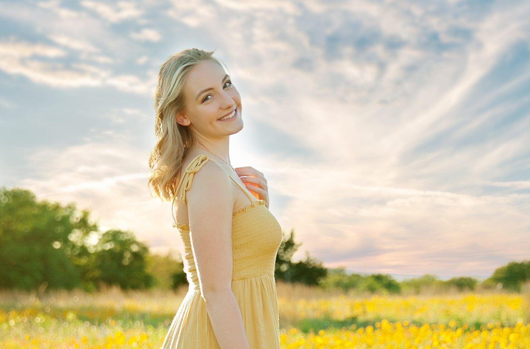 High school senior girl in a flowing yellow dress standing in a field of wildflowers during golden hour, smiling over her shoulder with the sun casting a warm glow. The sky is filled with soft, wispy clouds, creating a dreamy and radiant backdrop