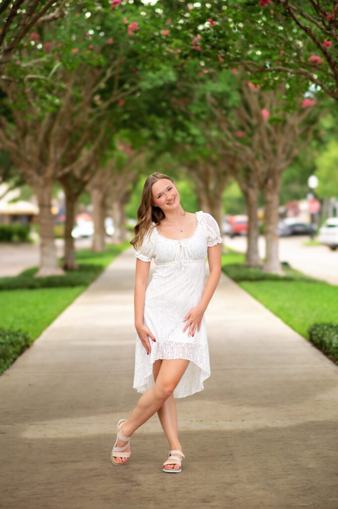 Senior girl in a white lace dress smiling under a tree-lined walkway in downtown Winter Garden during her senior photo session