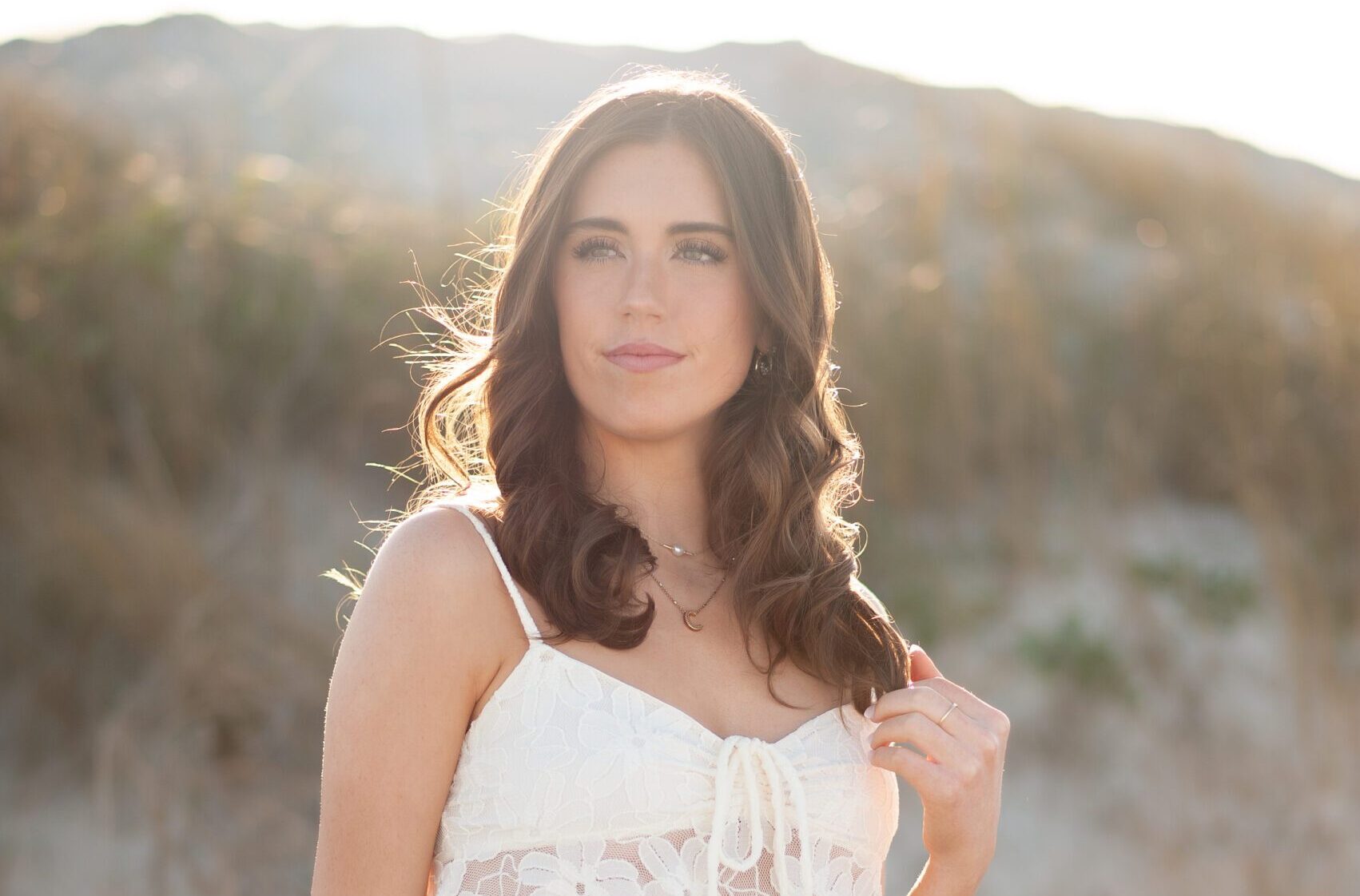 A young woman with wavy brown hair poses outdoors in soft, golden sunlight. She wears a delicate white lace crop top and blue jeans, gently touching her hair while gazing into the distance. The warm, backlit glow highlights her features, creating a dreamy and natural portrait. The blurred background of sand dunes and foliage adds to the serene, sun-kissed aesthetic. Orlando, FL