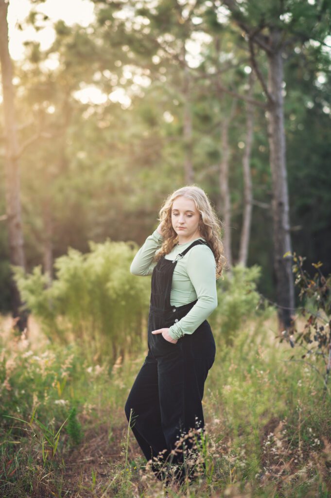 Senior posing casually in a soft green field near Orlando, FL wearing a sage top and overalls for her senior pictures