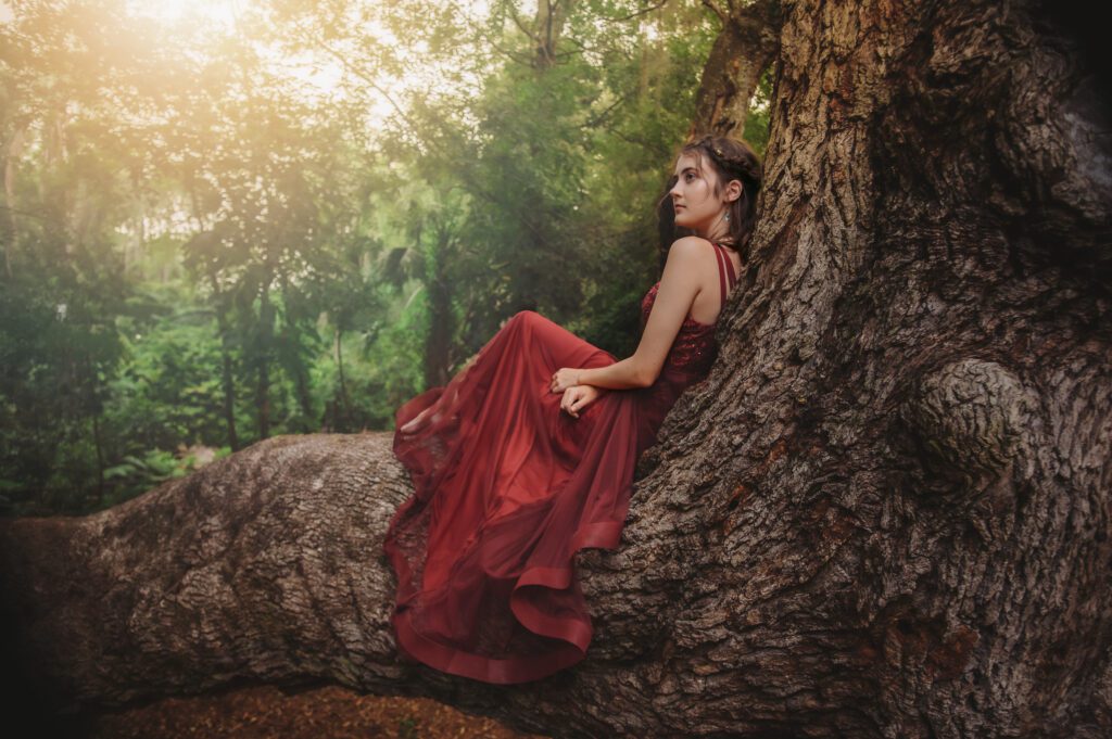 Senior sitting at the base of a large tree looking off with a relaxed expression during a whimsical golden hour shoot