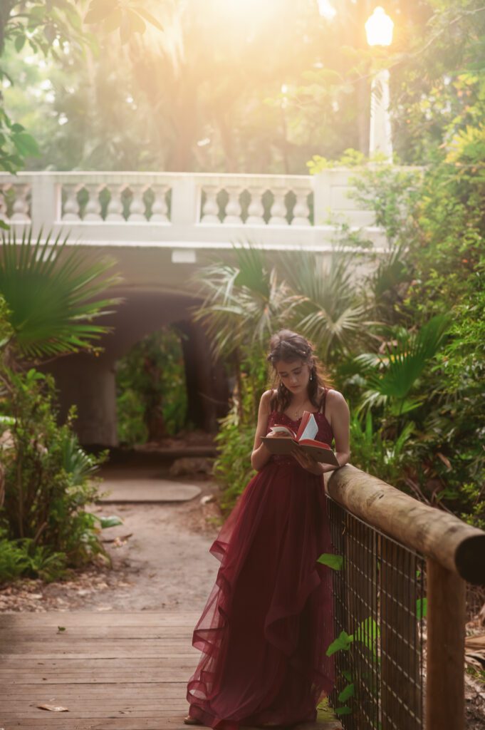 High school senior standing on a wooden bridge in a hidden park in Winter Park, reading a book in soft natural light