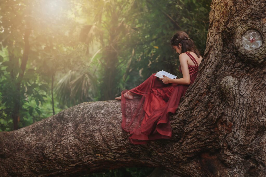 Senior sitting at the base of a large tree, reading a book during a whimsical golden hour shoot