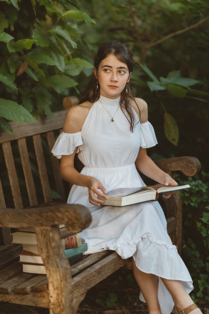 Senior girl in white dress sitting a bench looking through books in a park in Winter Park, FL near Orlando during her senior photo session with Orlando Senior Photographer, Melissa Vinsik.