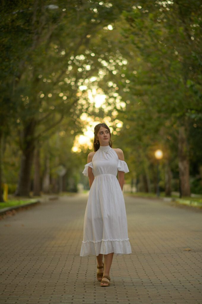 Senior girl in a white dress walking down road lined by trees during her senior photo session in a park near Orlando. 