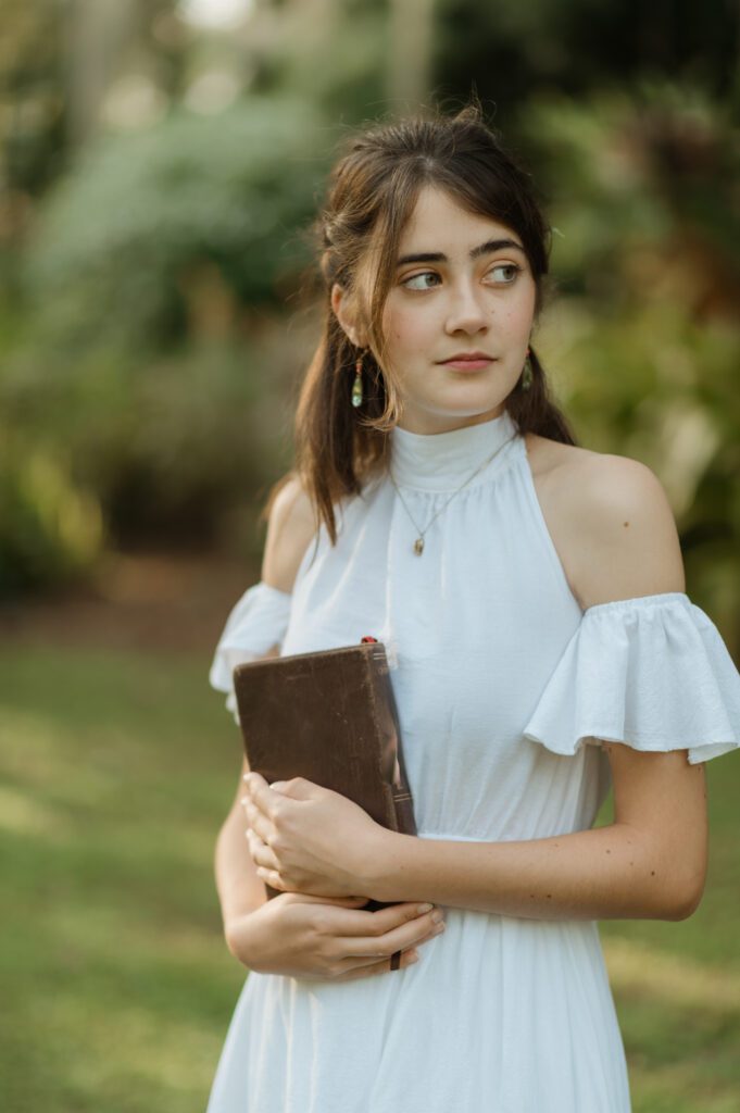 high school senior girl in a white dress holding her bible, glancing to the side during her senior photography session in a park near Orlando