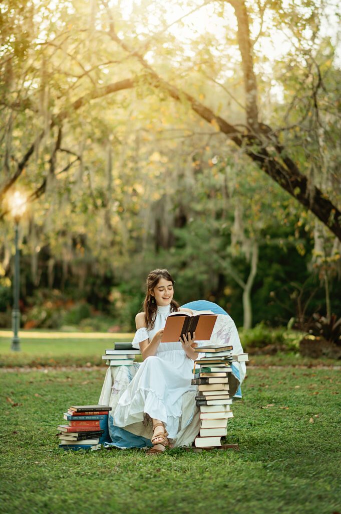 senior girl in white dress reading and smiling in a chair surrounded by books in Winter Park, Florida during her senior photo session with Melissa Vinsik Photography