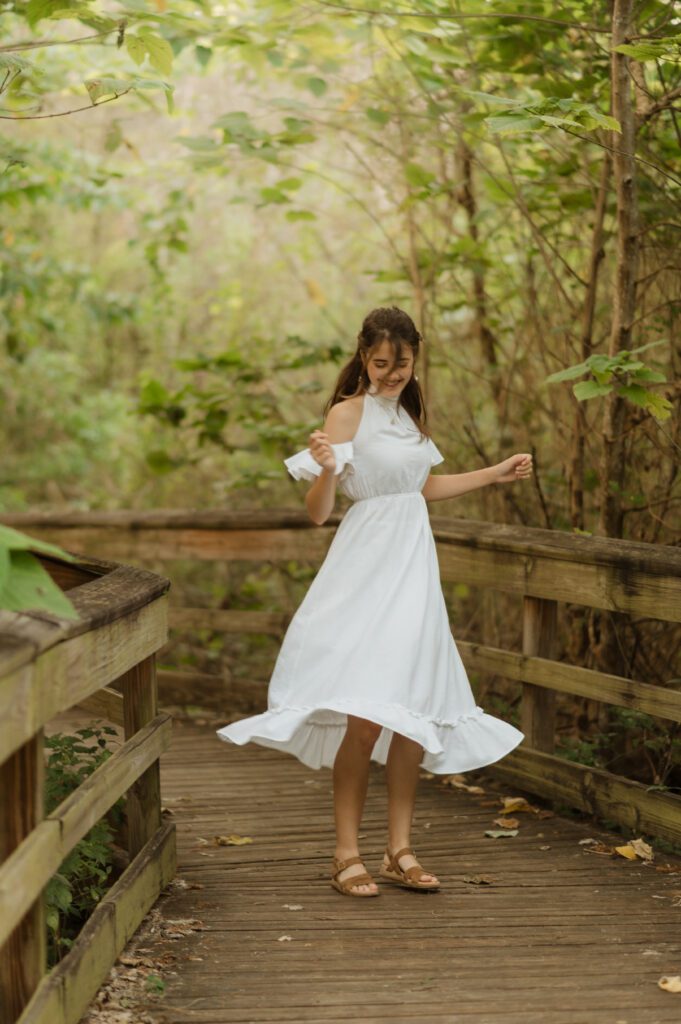 senior high school in white dress dancing on a boardwalk surrounded by greenery on a boardwalk at Mead Gardens in Winter Park, during her senior photo session with Melissa Vinsik Photography