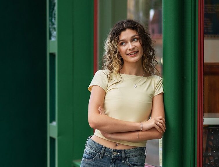 Senior girl smiling beside the green CityArts exterior in jeans and a yellow top.