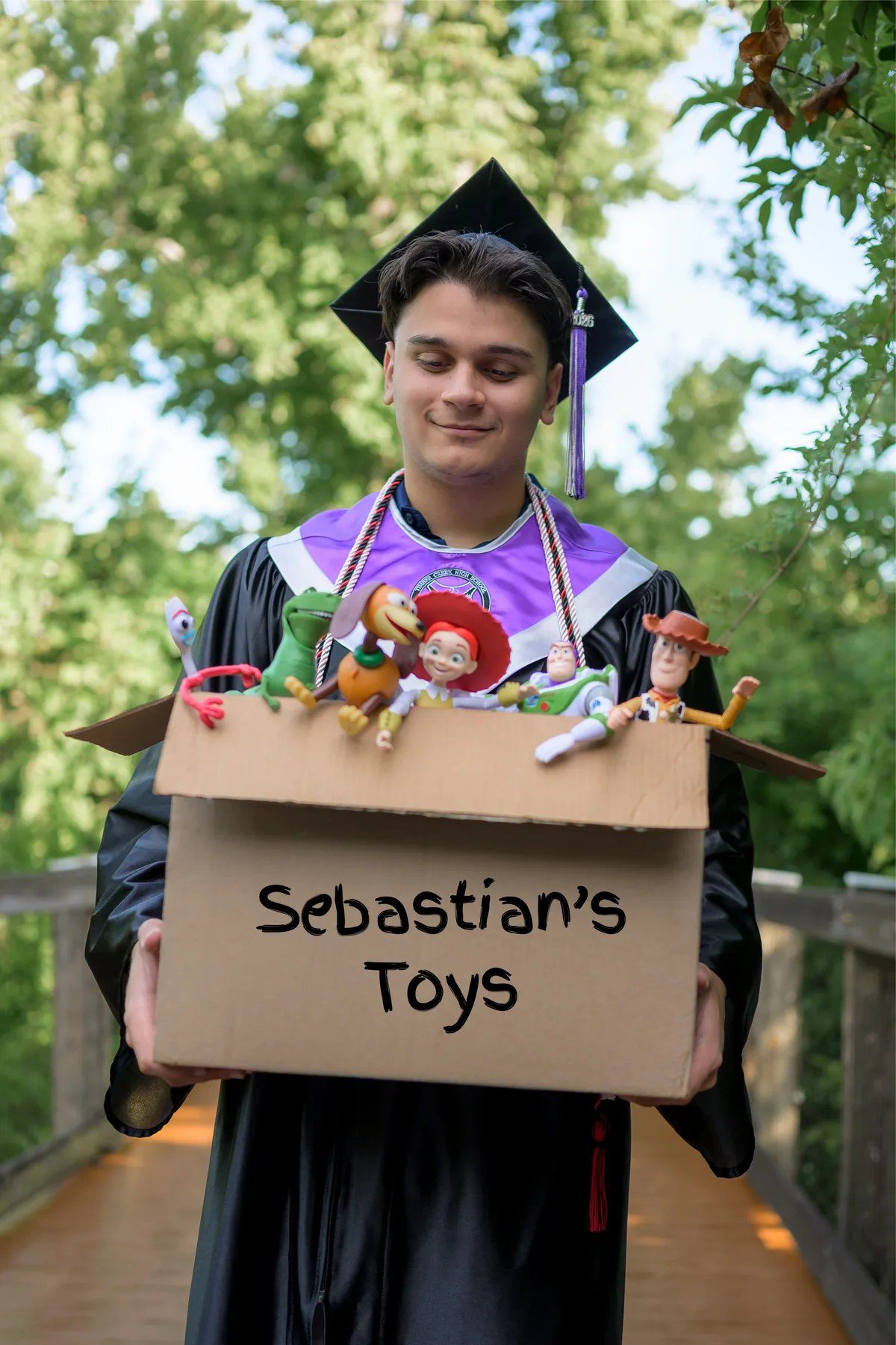 Toy Story-inspired graduation photo with a cap-and-gown senior holding a box of toys at Oakland Nature Preserve
