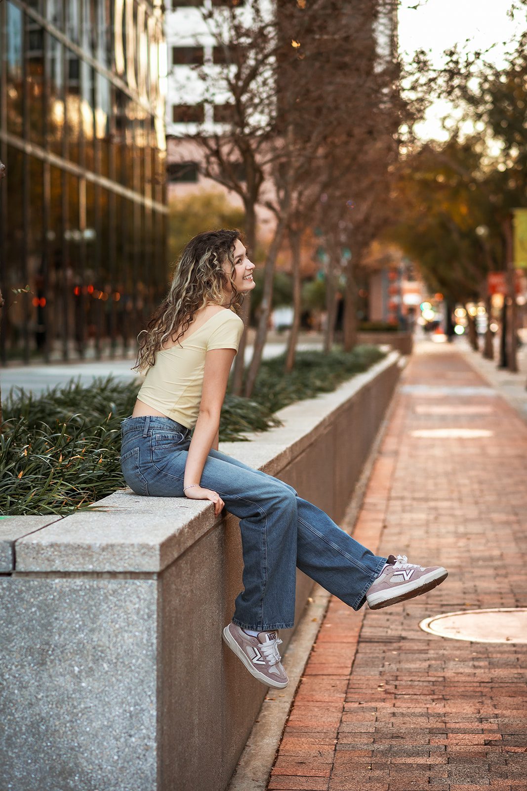 High school senior sitting on a stone wall with a brick sidewalk and city buildings behind her.