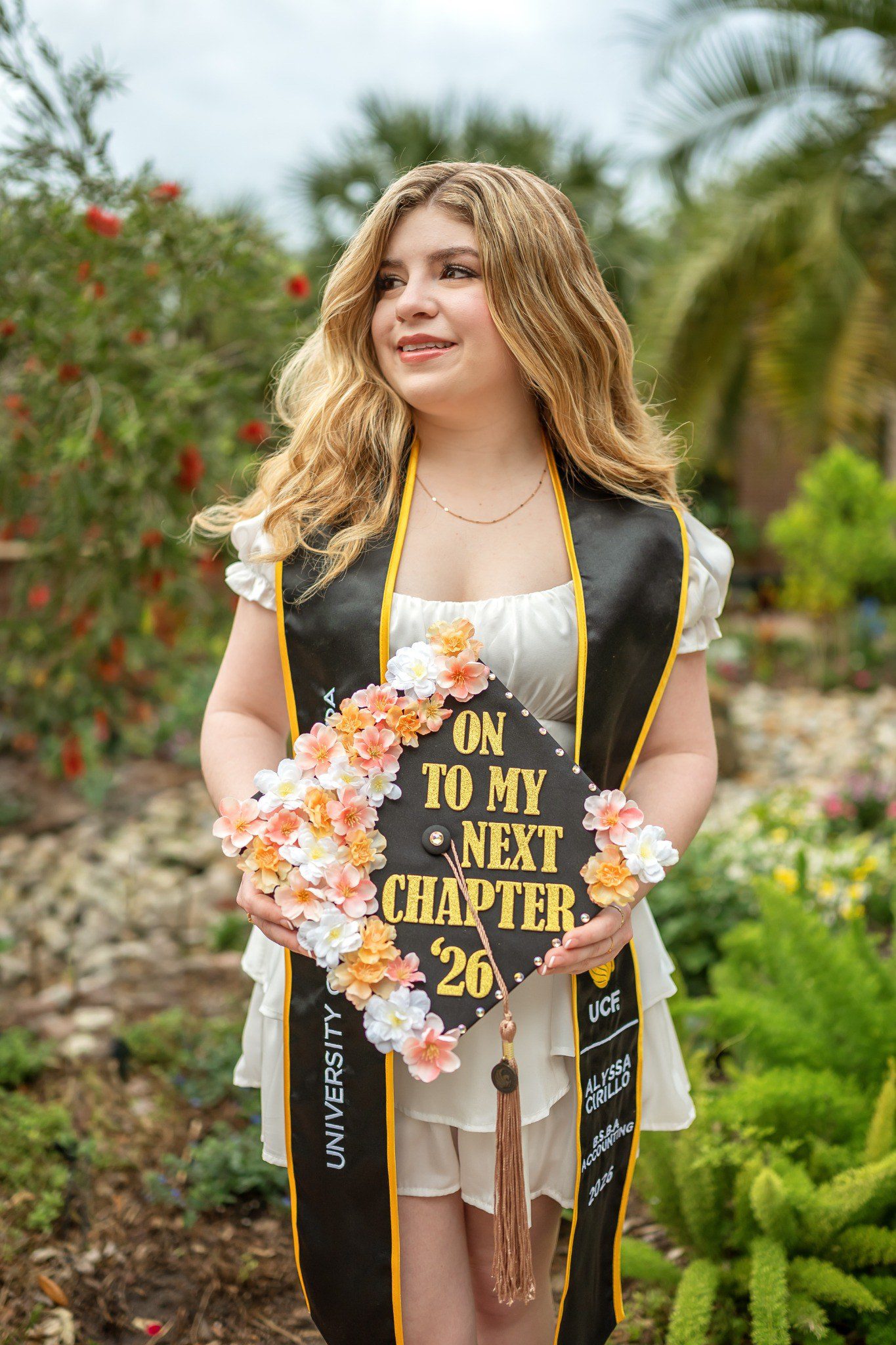 Floral graduation cap reading “On to my next chapter ’26” held against a black-and-gold stole.