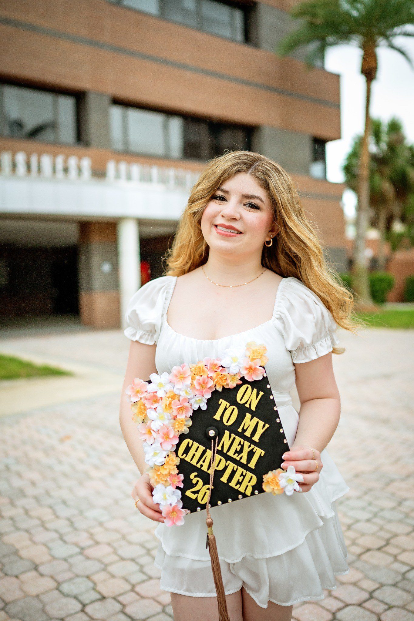 Close portrait of a graduate holding a flower-trimmed cap in front of the Business Administration building.