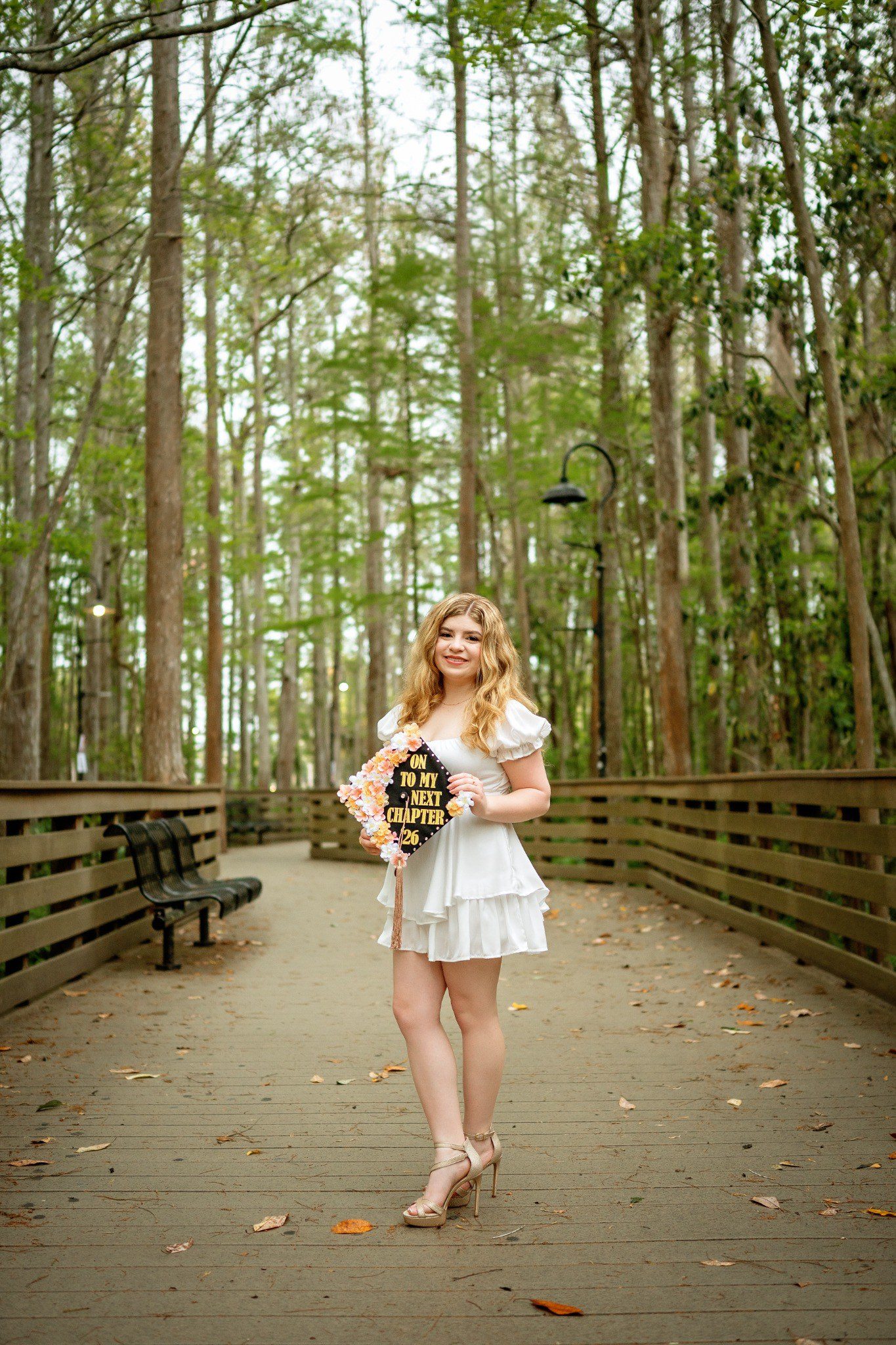 Graduate holding her decorated cap on a boardwalk surrounded by tall trees.