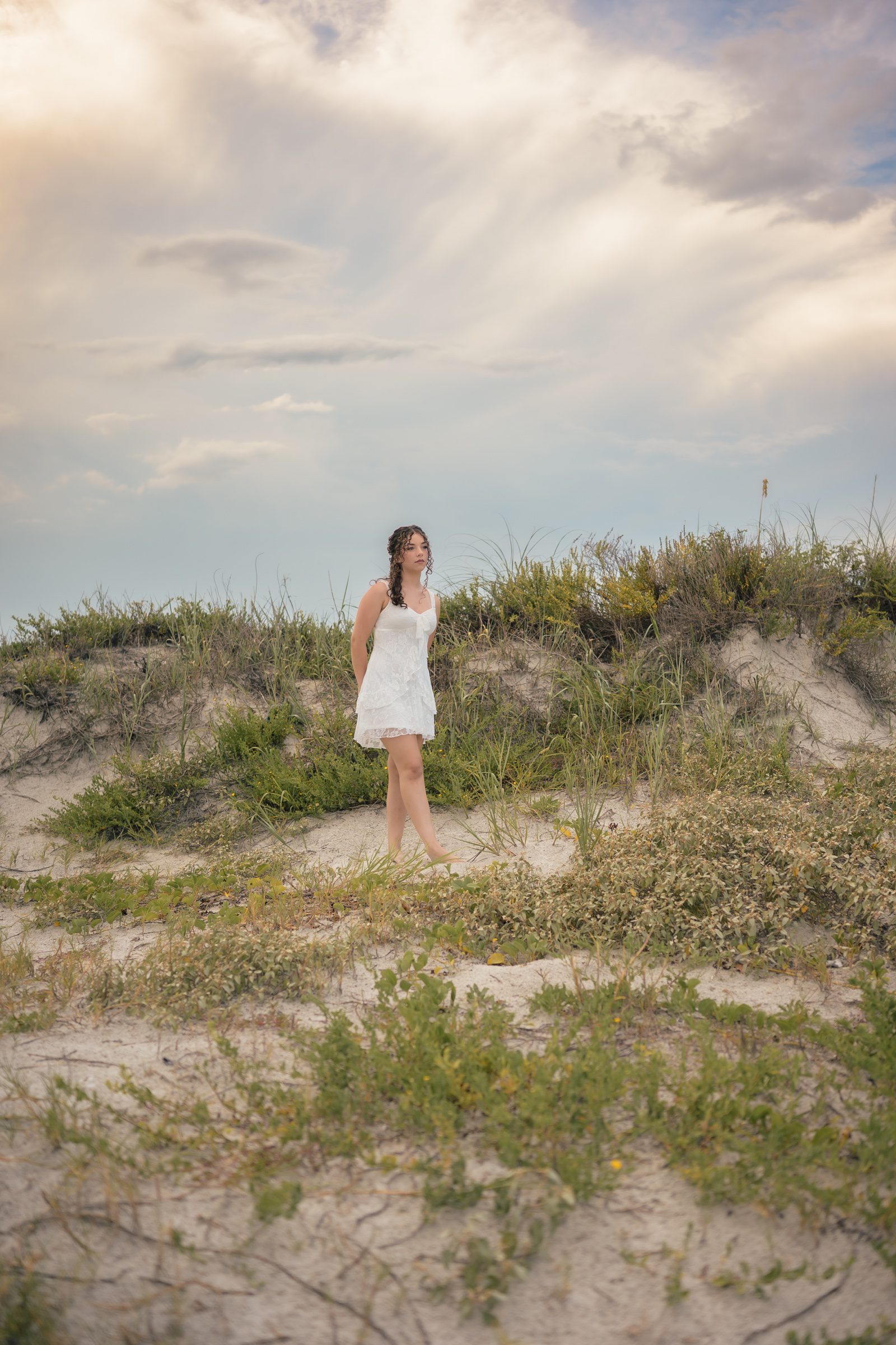 Senior in a white dress beside the dunes beneath a cloudy evening sky