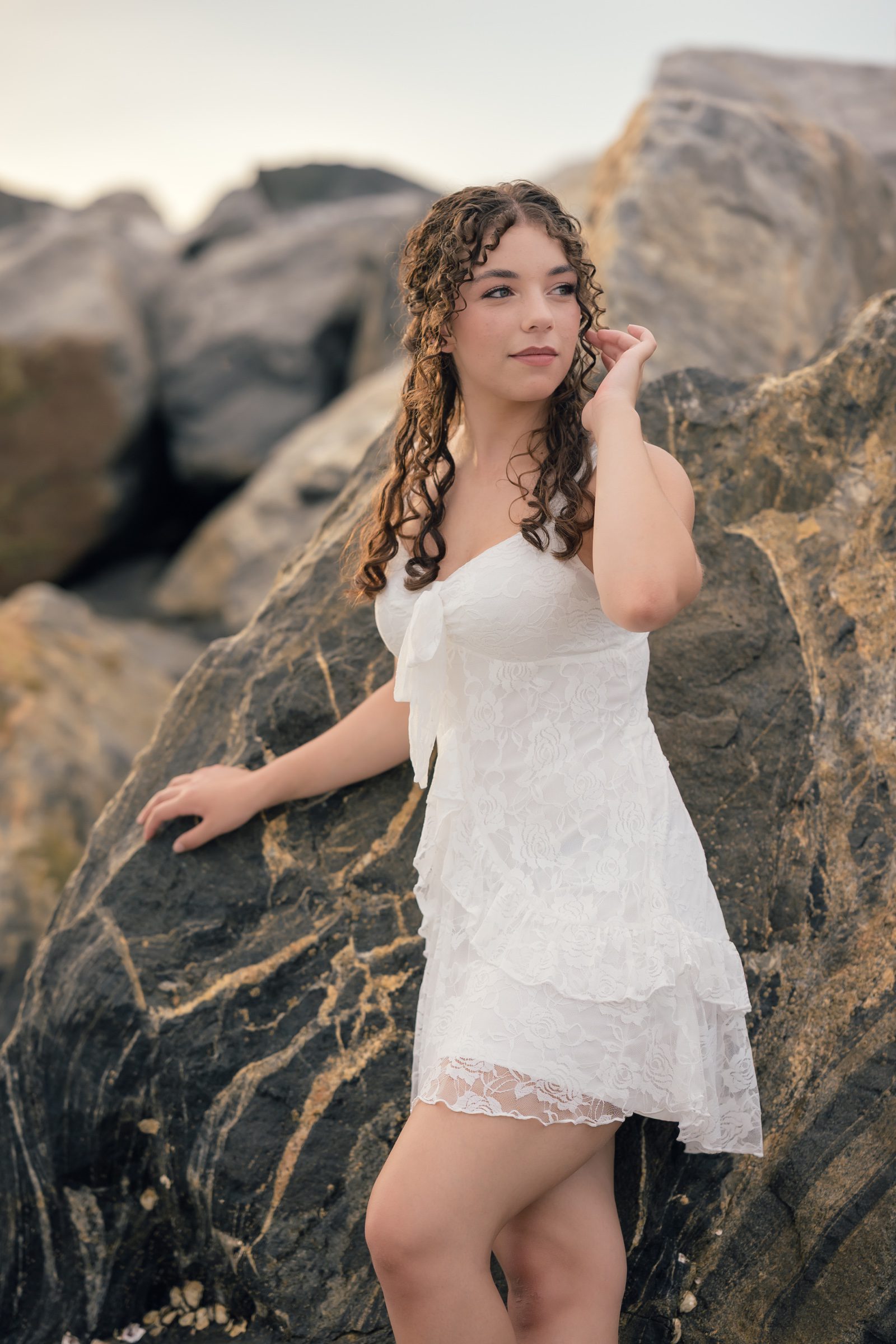 Senior posing in a white lace dress against the jetty rocks