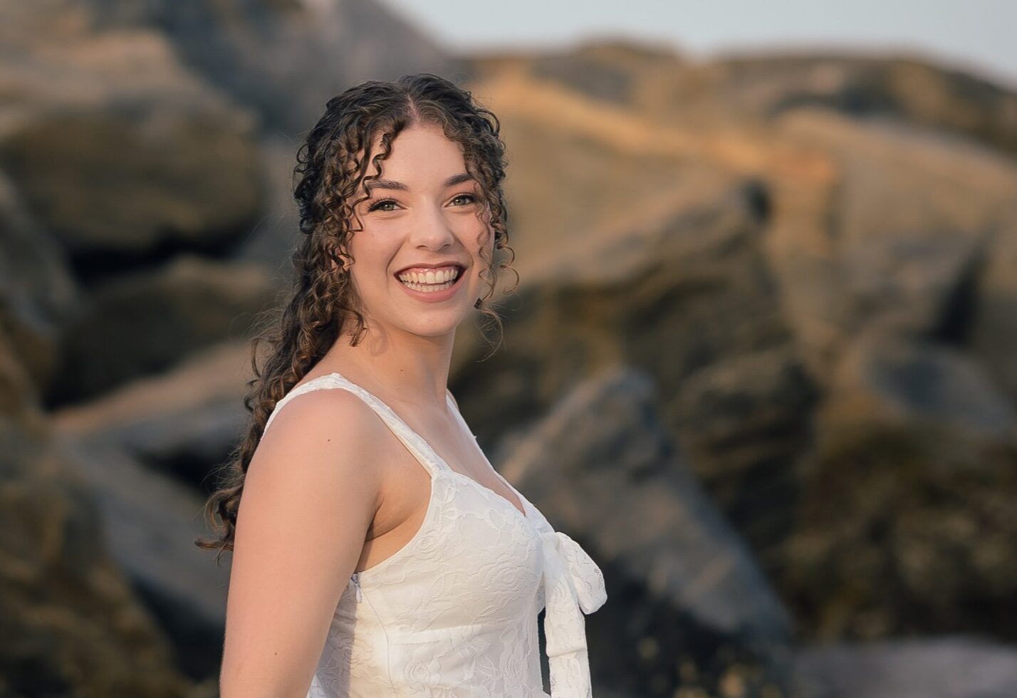Senior smiling in a white dress beside the water and rocks