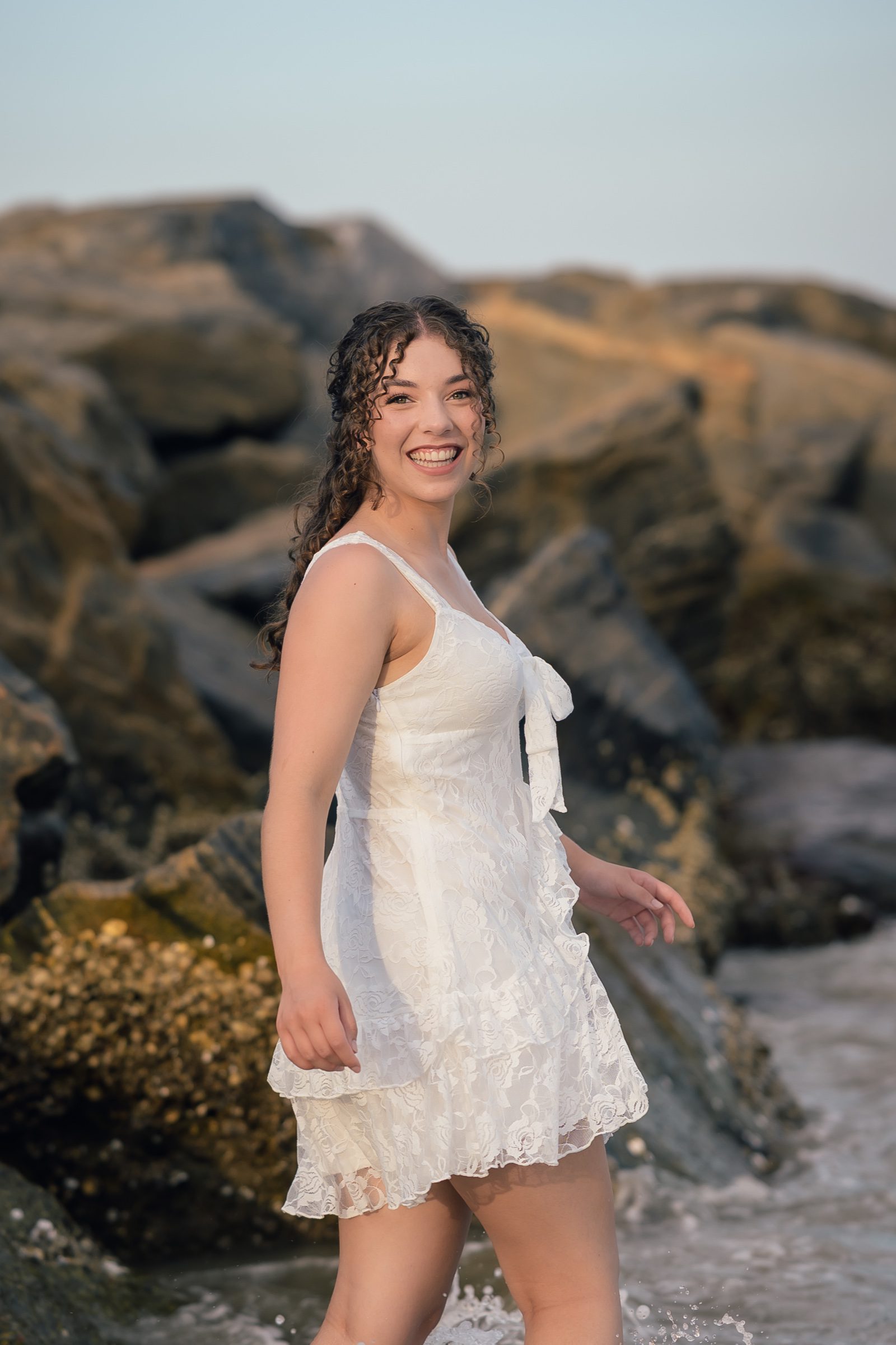 Senior smiling in a white dress beside the water and rocks