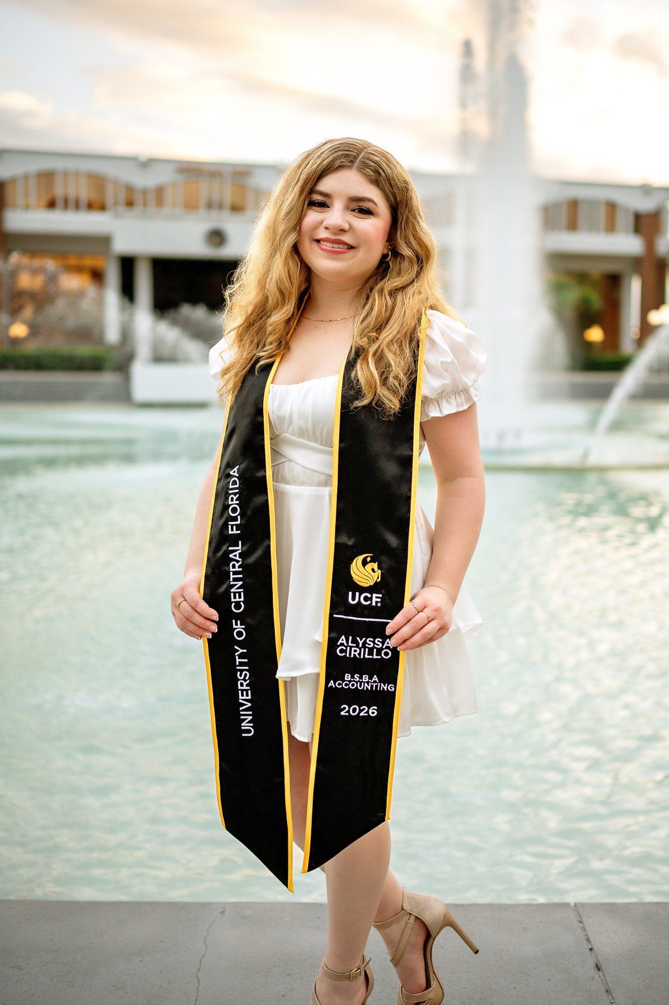Graduate wearing a personalized UCF stole marked B.S.B.A. Accounting beside the campus fountain.