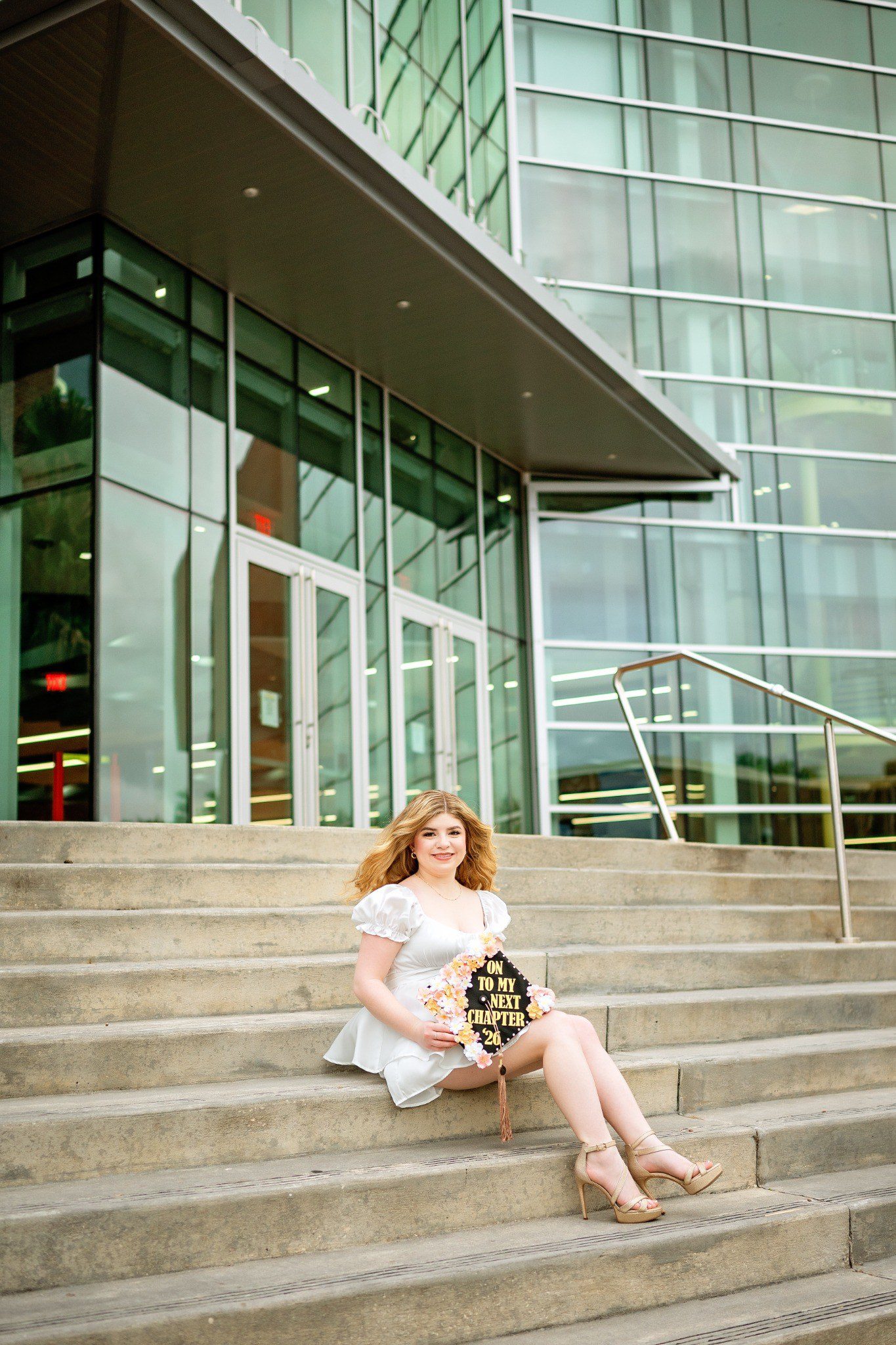 Graduate seated on campus steps in a white outfit with her decorated cap beside her.