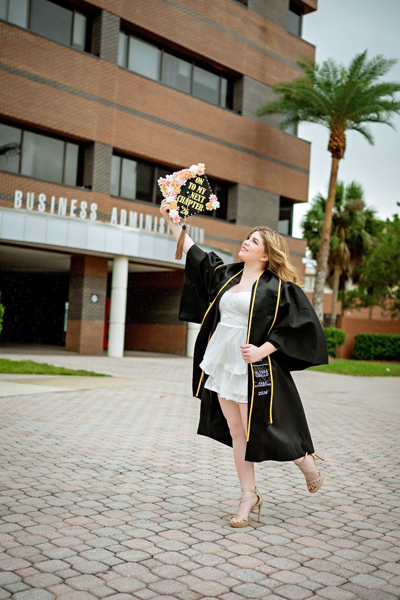 Graduate lifting her cap and bending one leg outside the Business Administration building.
