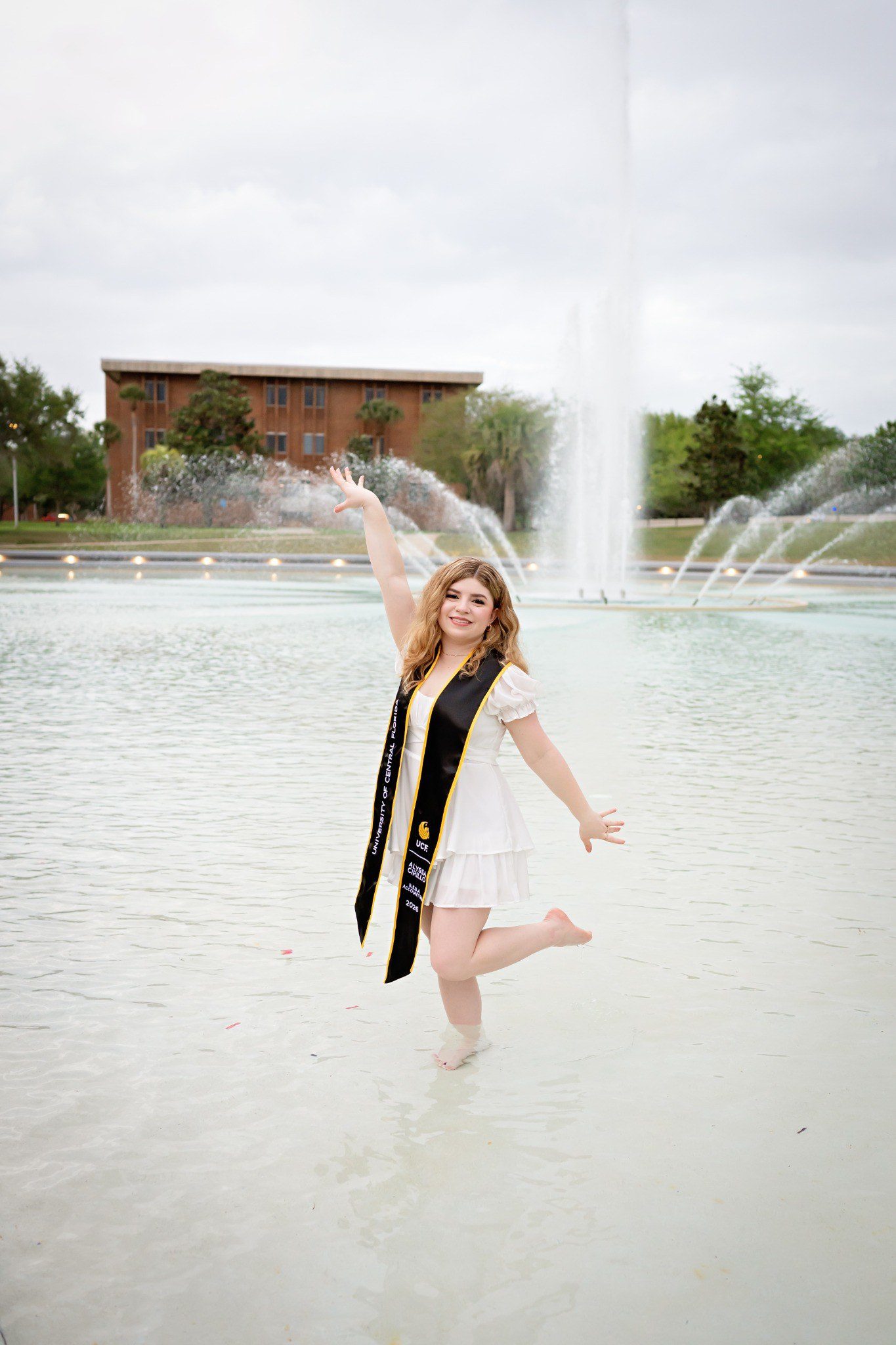 Graduate lifting one arm and bending one leg while standing in the campus fountain.