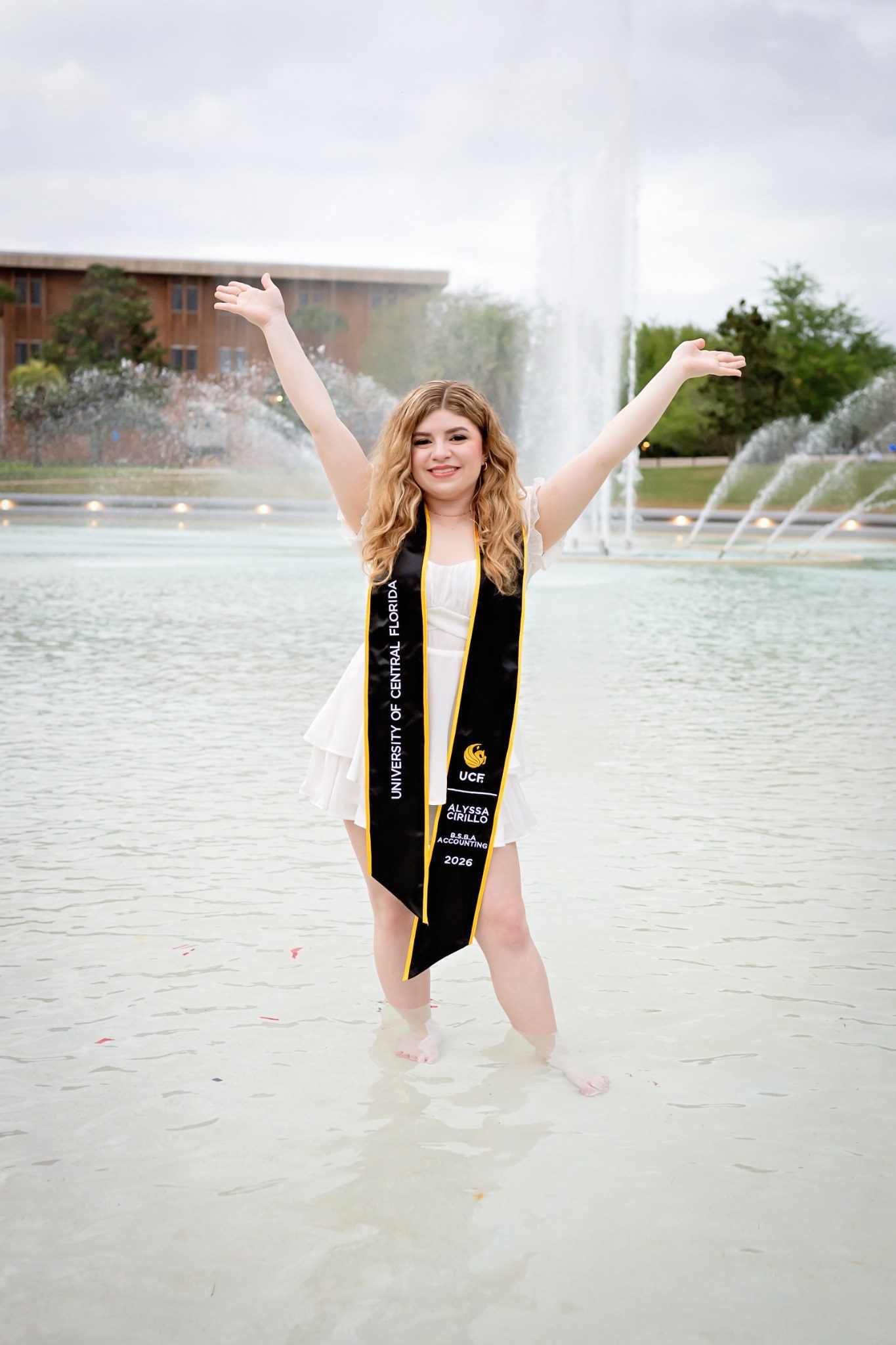 Graduate raising both arms in celebration while standing in UCF’s fountain.