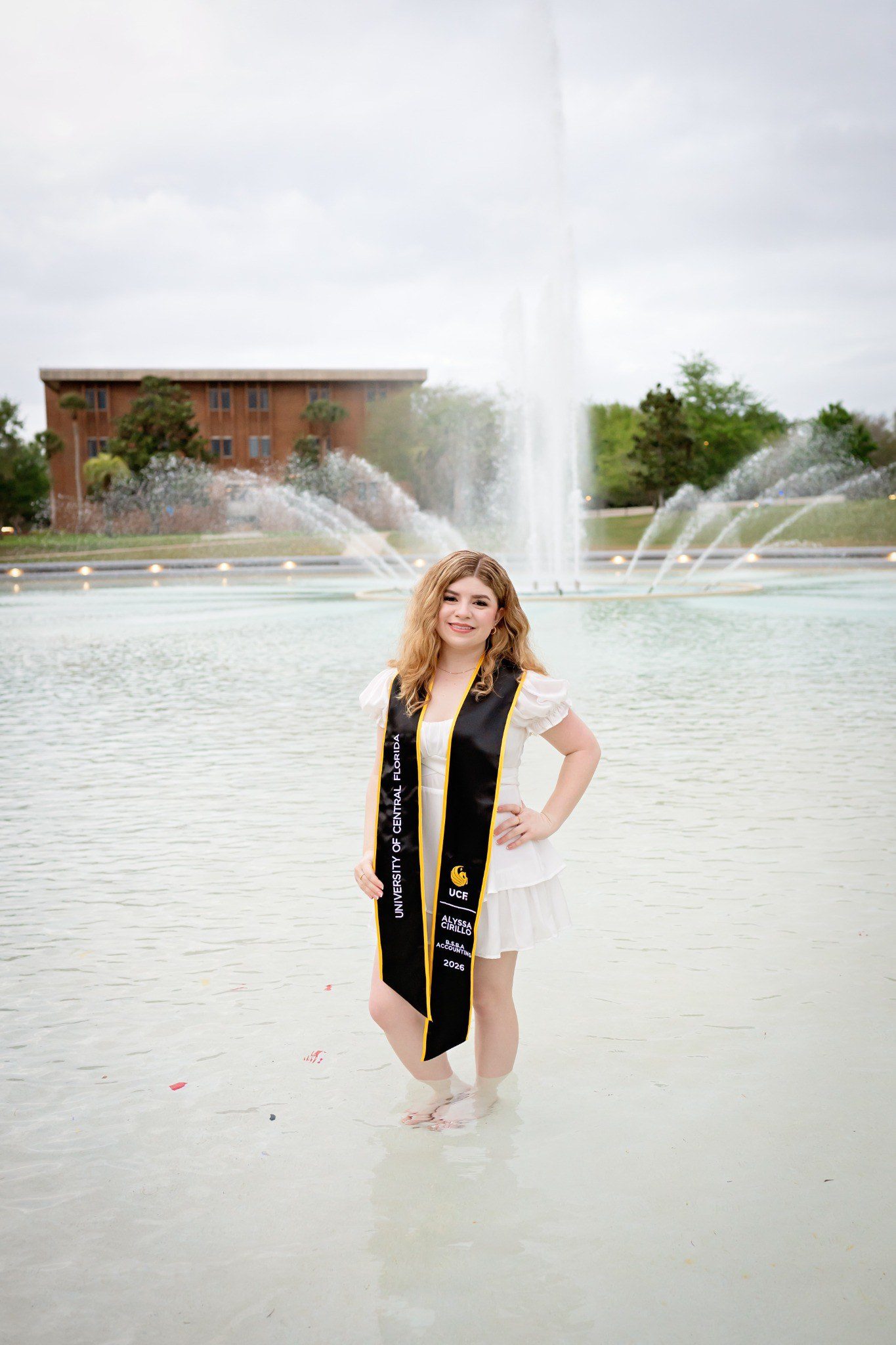Graduate posing with one hand on her hip in the fountain, wearing a black-and-gold UCF stole.