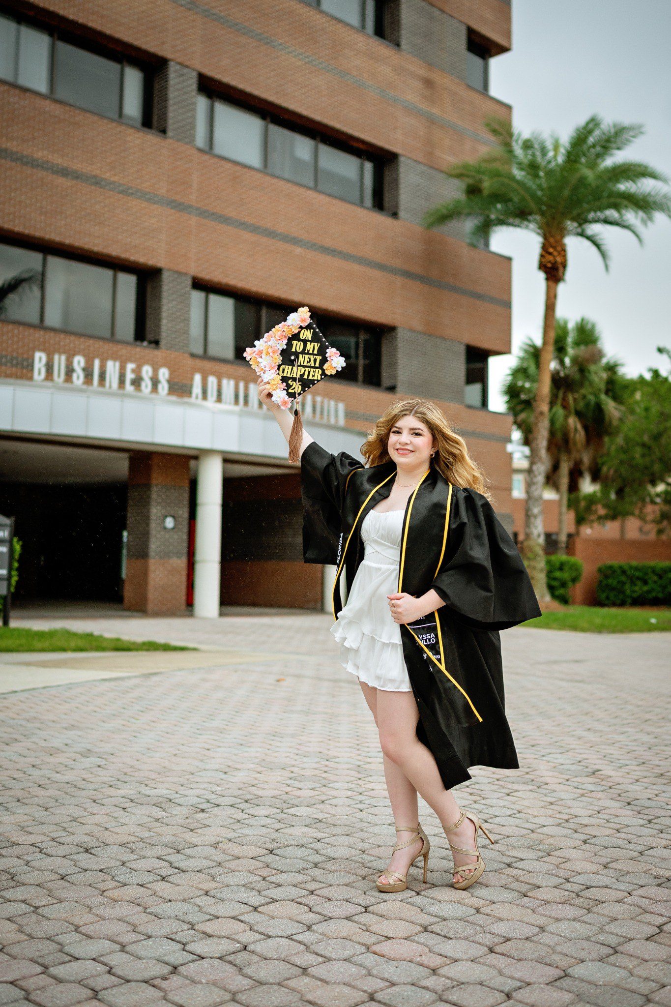 Graduate in a black gown raising her decorated cap outside UCF’s Business Administration building.