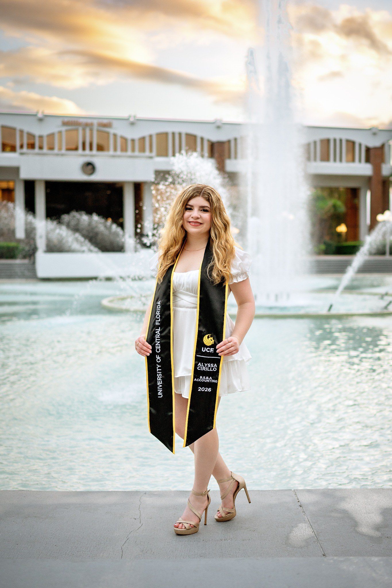Full-length graduation portrait beside UCF’s fountain with a personalized black-and-gold stole.