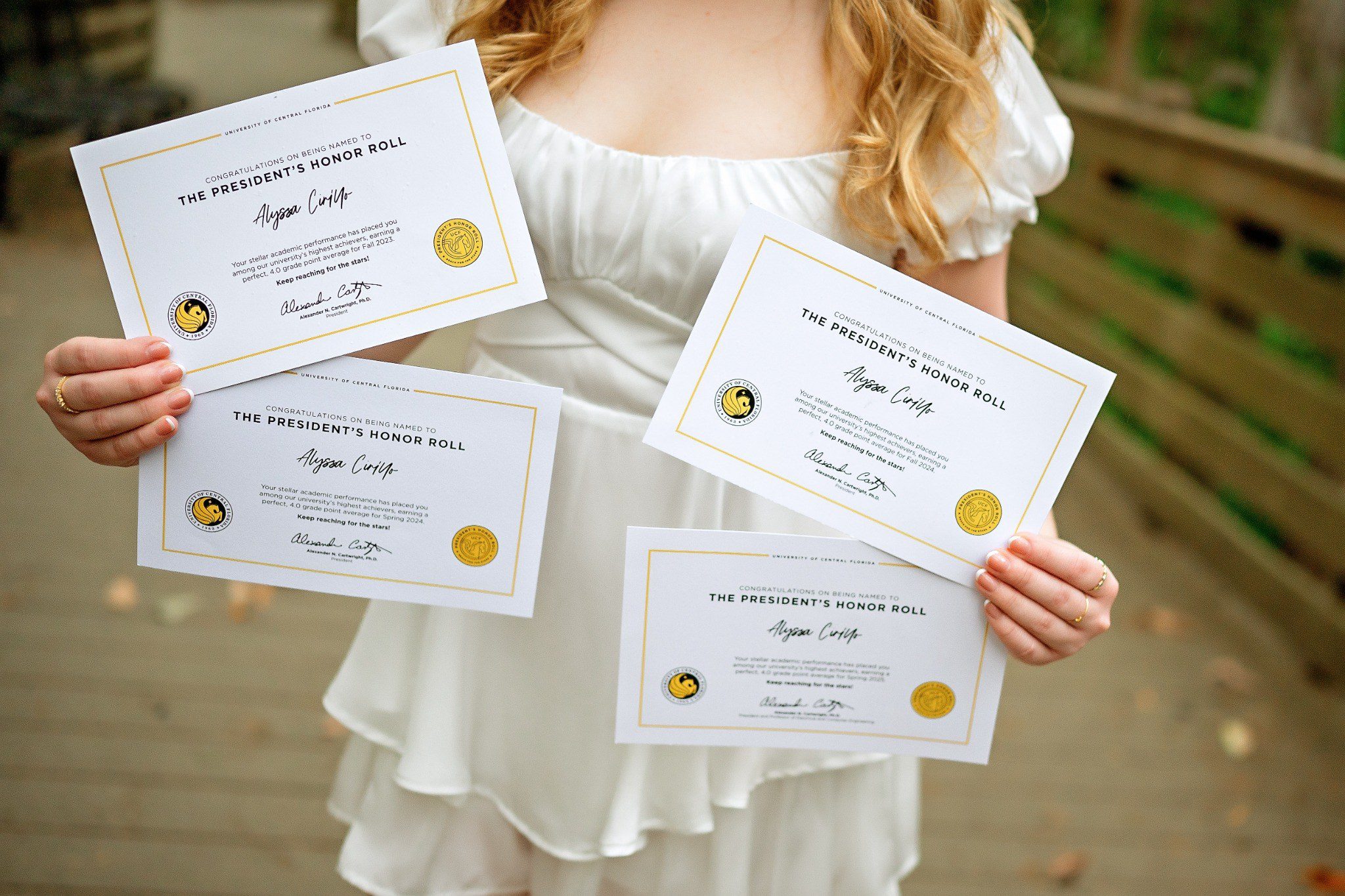 Graduate holding four President’s Honor Roll certificates in front of her white outfit.