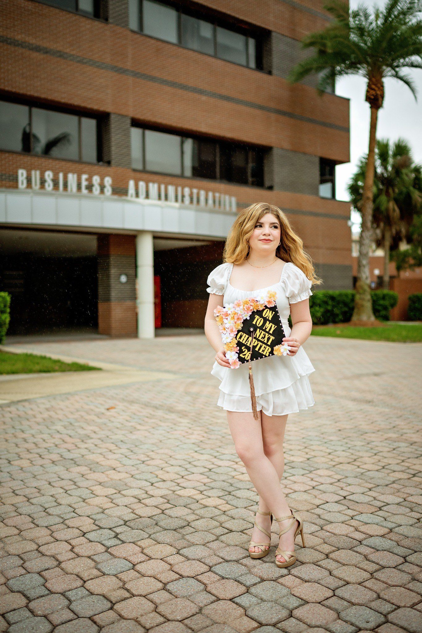 Graduate in a white outfit holding her decorated cap outside UCF’s Business Administration building.