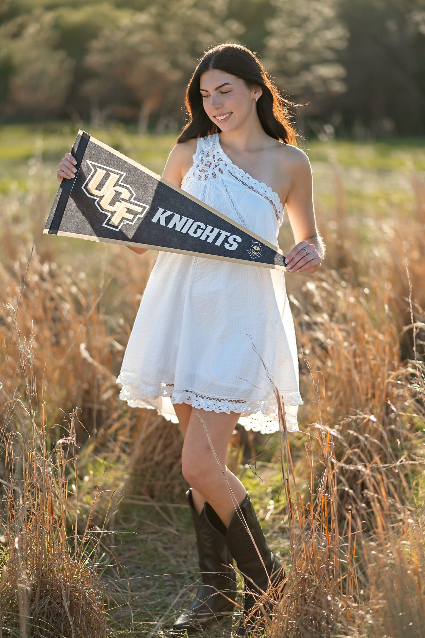 Senior holding a UCF Knights pennant in a white dress in a Lake Louisa field