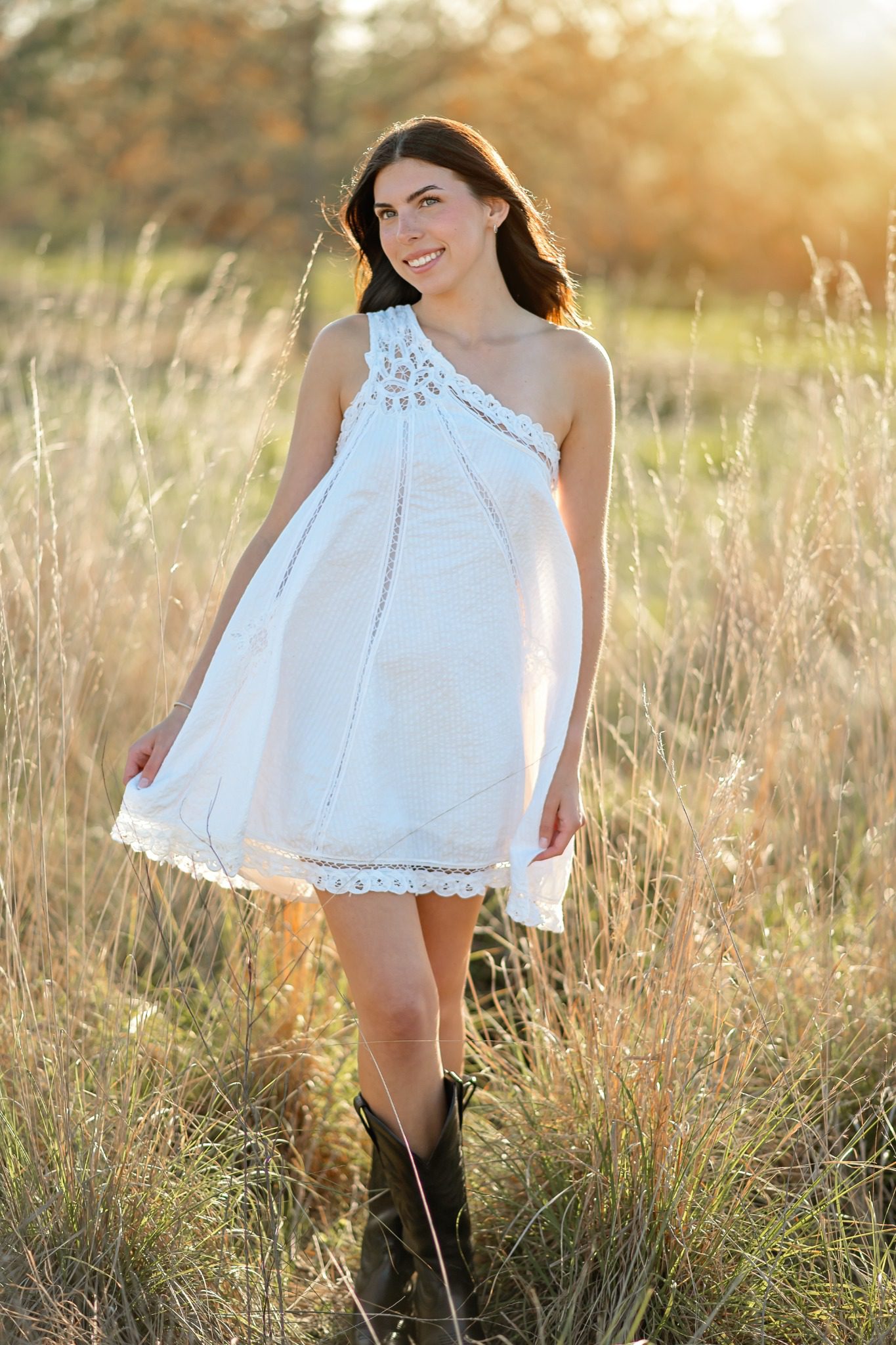 Senior holding the hem of a white dress in a sunlit field at Lake Louisa