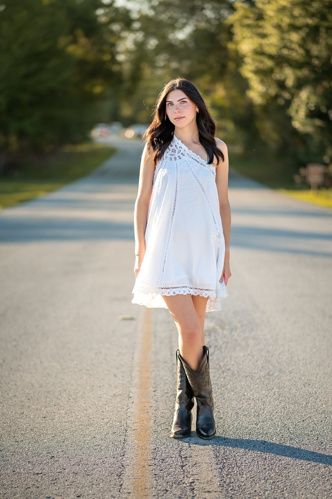 Full-length senior portrait in a white dress and black boots on a tree-lined park road
