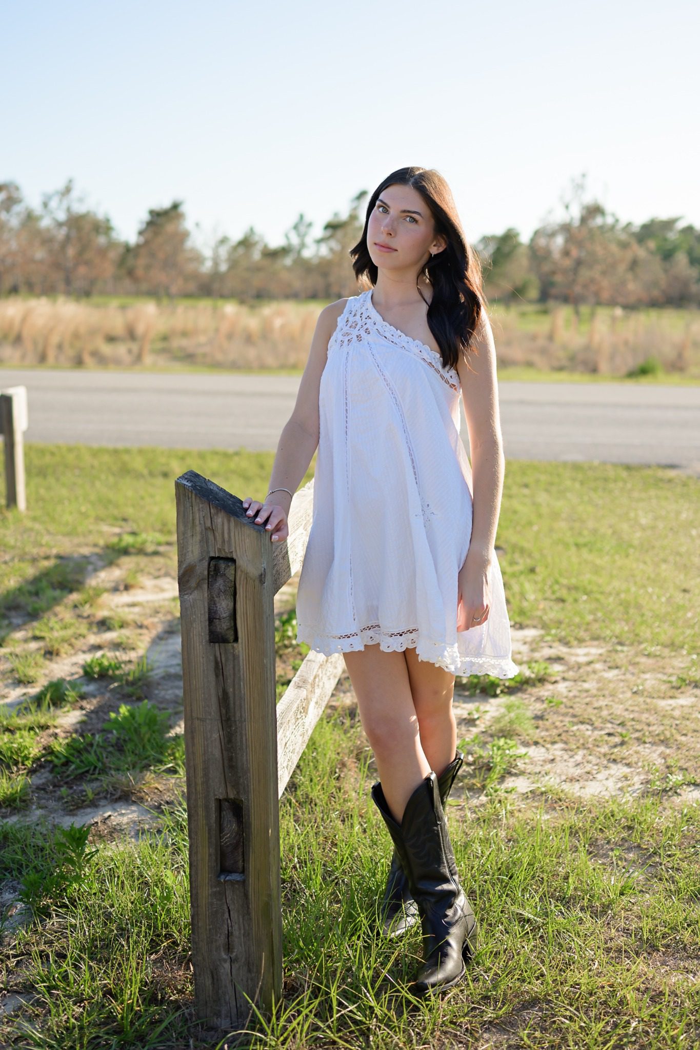 Senior in a white dress and black boots leaning beside a wooden fence at Lake Louisa